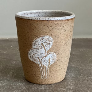 Op de afbeelding: Een bruine keramische mok met een gespikkelde glazuur en een wit ontwerp van drie paddenstoelen.