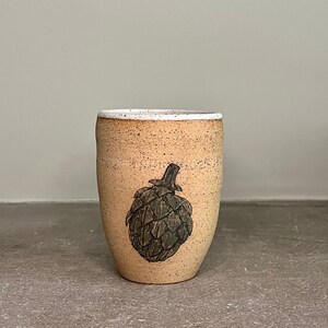 May include: A beige ceramic mug with a white rim and a black and green illustration of an artichoke.