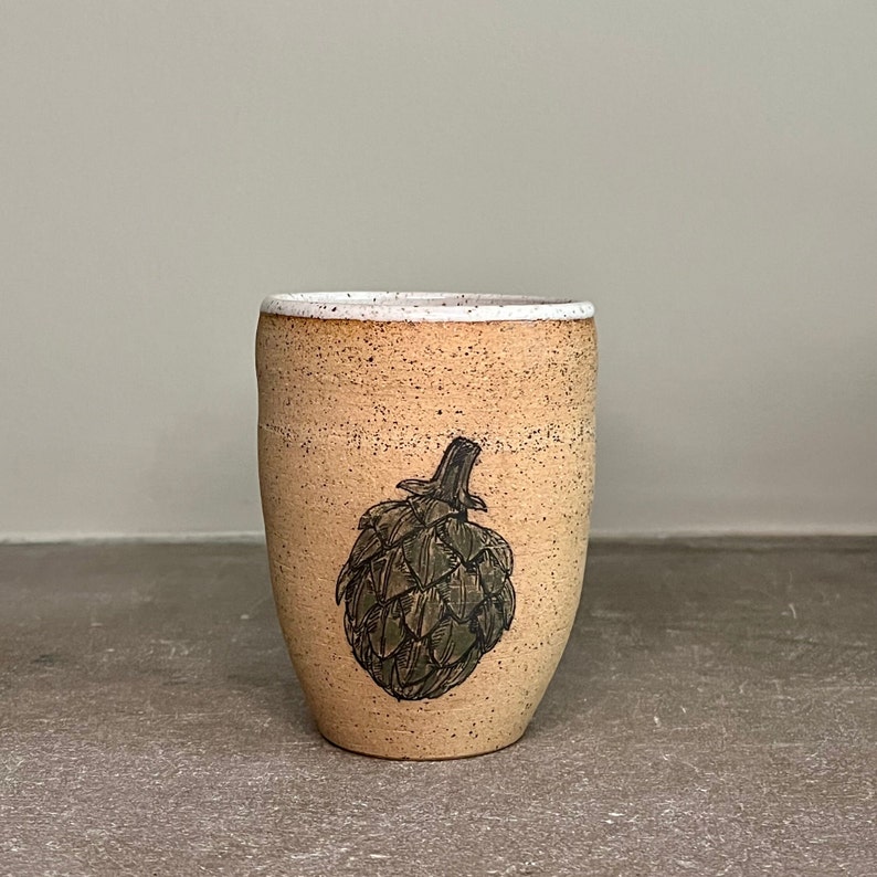May include: A beige ceramic mug with a white rim and a black and green illustration of an artichoke.