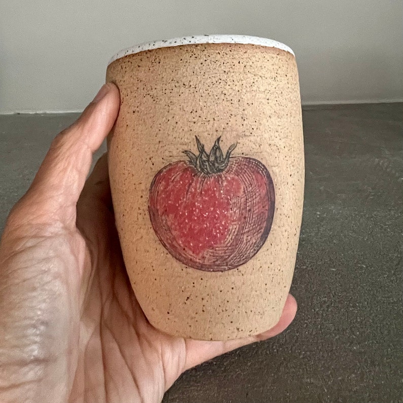 May include: A beige ceramic mug with a speckled glaze and a white rim. The mug features a hand-drawn illustration of a red tomato with a green stem.