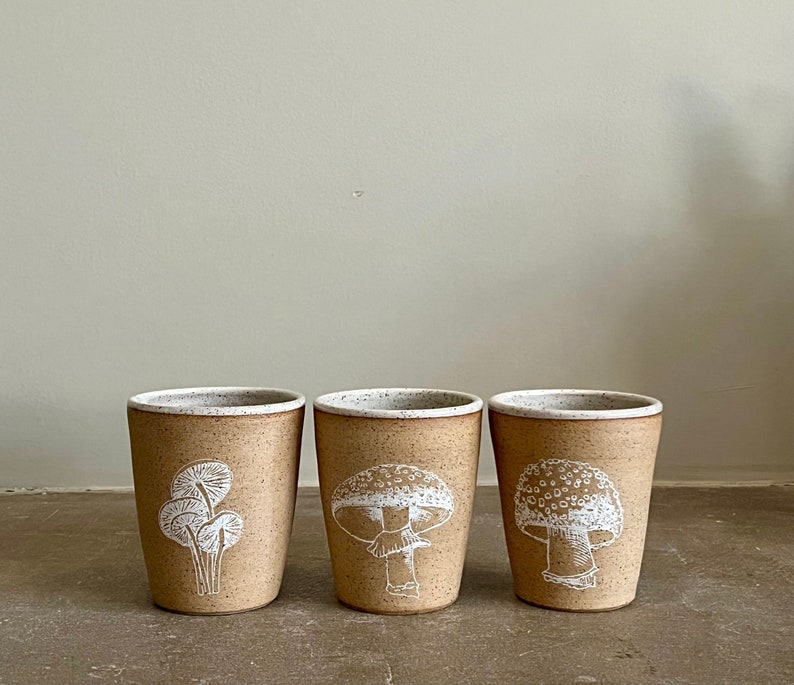 Op de afbeelding: Drie keramische mokken met witte illustraties van paddenstoelen en varens. De mokken zijn lichtbruin van kleur met een gespikkeld wit glazuur.