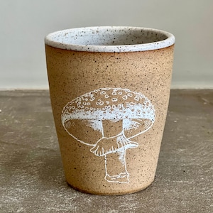 Op de afbeelding: Een beige keramische mok met een wit gespikkeld glazuur. De mok heeft een witte illustratie van een paddenstoel met een wit gespikkeld hoedje.