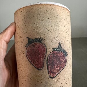 May include: A beige ceramic mug with a speckled glaze and two hand-painted strawberries in red and black.