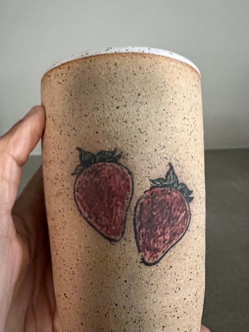 May include: A beige ceramic mug with a speckled glaze and two hand-painted strawberries in red and black.