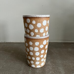 May include: Two brown ceramic mugs with white polka dots. The mugs are stacked on top of each other.