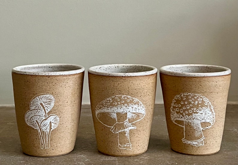 Op de afbeelding: Drie keramische mokken met witte paddenstoel illustraties. De mokken zijn bruin met een gespikkeld wit glazuur.