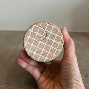 Op de afbeelding: Een ronde, bruine keramische pot met een wit rasterpatroon. De pot heeft een gat in het midden voor drainage.