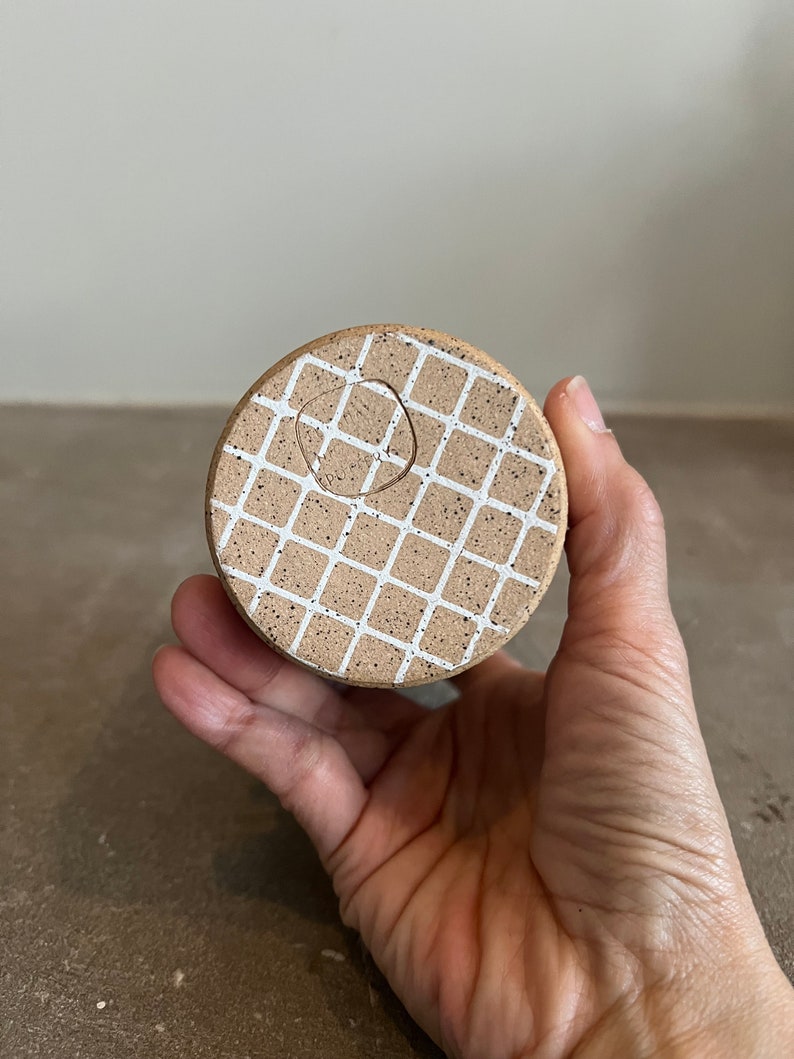 Op de afbeelding: Een ronde, bruine keramische pot met een wit rasterpatroon. De pot heeft een gat in het midden voor drainage.