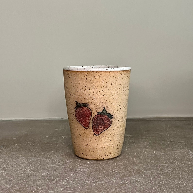May include: A beige ceramic mug with a speckled glaze and two hand-drawn strawberries in red and green.
