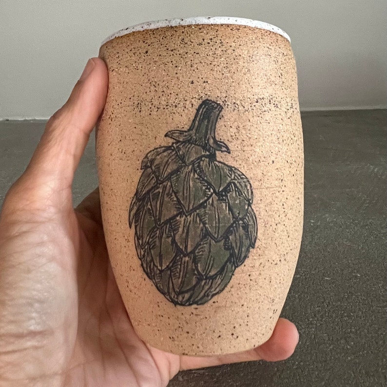 May include: A beige ceramic mug with a white rim and a black and white illustration of an artichoke.