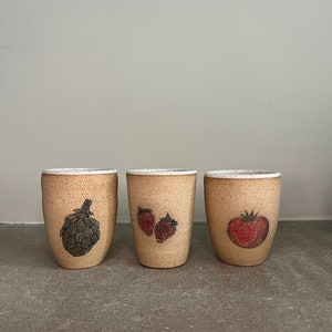 May include: Three ceramic mugs with different vegetable illustrations. The mug on the left has an artichoke, the middle mug has two strawberries, and the mug on the right has a tomato.