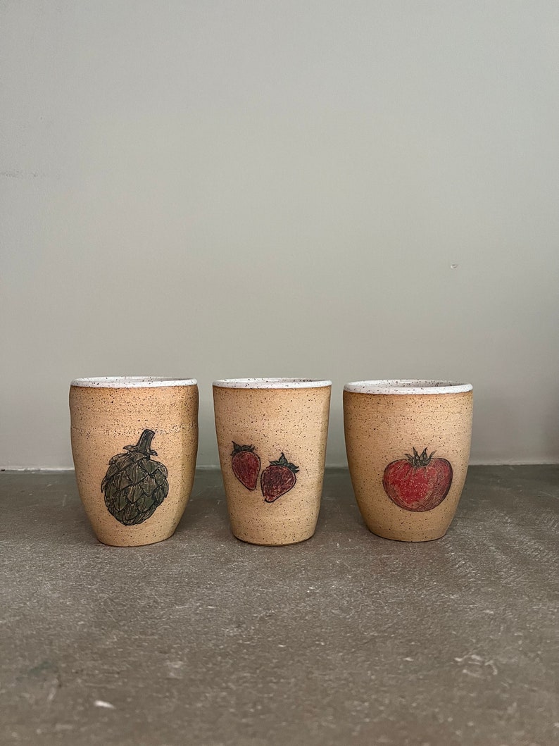 May include: Three ceramic mugs with different vegetable illustrations. The mug on the left has an artichoke, the middle mug has two strawberries, and the mug on the right has a tomato.
