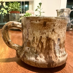 May include: A handmade ceramic mug with a textured, earthy appearance. The mug features a brown and cream color palette with a unique, organic design. The handle is integrated into the mug's form, creating a cohesive look. The mug is suitable for hot or cold beverages.