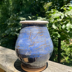 May include: A handmade ceramic vase with a blue and brown glaze. The vase has a wide body and a narrow neck with a white rim. The base is a light brown color. The vase is sitting on a wooden surface.