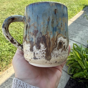 May include: A ceramic mug with a rounded body and a curved handle. The upper portion features a glossy blue glaze that transitions into a dark brown drip pattern over a speckled cream-colored base. The surface has a textured, handmade appearance.