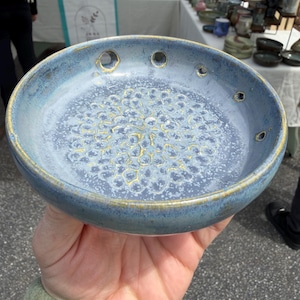 Pode incluir: Uma tigela de cer&acirc;mica redonda com esmalte salpicado em azul e branco e um padr&atilde;o texturizado em tom dourado no centro. A borda apresenta v&aacute;rios pequenos orif&iacute;cios circulares espa&ccedil;ados e o rebordo exterior tem um acabamento dourado.