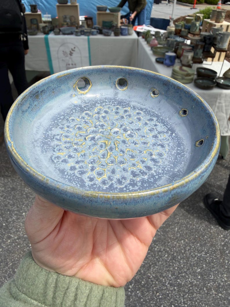 Pode incluir: Uma tigela de cer&acirc;mica redonda com esmalte salpicado em azul e branco e um padr&atilde;o texturizado em tom dourado no centro. A borda apresenta v&aacute;rios pequenos orif&iacute;cios circulares espa&ccedil;ados e o rebordo exterior tem um acabamento dourado.
