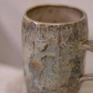 May include: A handmade ceramic mug with a textured surface and a handle. The mug features a blend of colors, including blue, brown, and gray, creating a unique, rustic appearance. The mug is designed for hot beverages.
