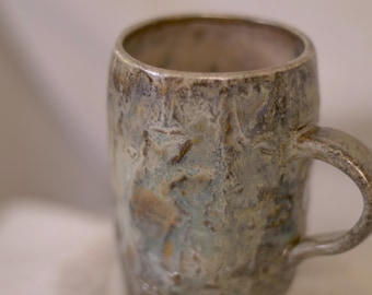 Pode incluir: Uma caneca de cerâmica artesanal com uma superfície texturizada e uma alça. A caneca apresenta uma mistura de cores, incluindo azul, castanho e cinzento, criando uma aparência rústica única. A caneca é projetada para bebidas quentes.