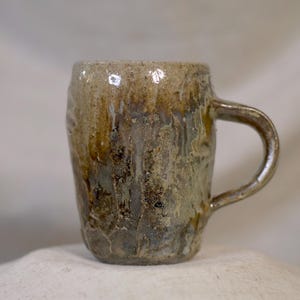 May include: A handmade ceramic mug with a textured surface and a handle. The mug features a blend of earthy tones, including brown, tan, and green, with a glossy finish. The mug is approximately 4 inches tall.