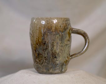 Pode incluir: Uma caneca de cerâmica artesanal com uma superfície texturizada e uma alça. A caneca apresenta uma mistura de tons terrosos, incluindo castanho, bege e verde, com um acabamento brilhante. A caneca tem cerca de 10 cm de altura.