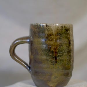 Puede incluir: Una taza de cerámica hecha a mano con una superficie texturizada única. La taza presenta una paleta de colores oscuros y terrosos con tonos de marrón, verde y dorado. El asa es suavemente curvada y la taza mide unos 10 cm de alto.