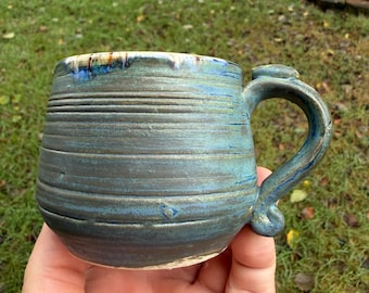 Pode incluir: Uma caneca de cerâmica feita à mão com corpo arredondado e alça curva. O exterior apresenta uma superfície texturizada com estrias horizontais, com acabamento em esmalte azul profundo e verde-azulado com toques de branco e castanho no rebordo.