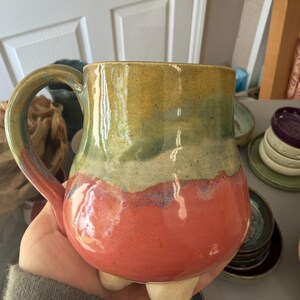 May include: Handmade ceramic mug with a unique color gradient. The mug transitions from a yellow-green top to a green middle, and a vibrant red base. It features a rounded body, a sturdy handle, and three small feet for stability. A piece of functional art.