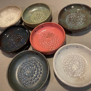 Pode incluir: Uma cole&ccedil;&atilde;o de seis pratos raladores de cer&acirc;mica circulares em v&aacute;rias cores, incluindo branco, verde s&aacute;lvia, castanho escuro, azul profundo, rosa coral e cinzento ard&oacute;sia. Cada prato apresenta um padr&atilde;o floral central texturizado.