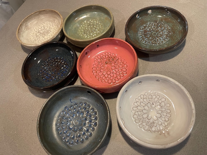 Pode incluir: Uma cole&ccedil;&atilde;o de seis pratos raladores de cer&acirc;mica circulares em v&aacute;rias cores, incluindo branco, verde s&aacute;lvia, castanho escuro, azul profundo, rosa coral e cinzento ard&oacute;sia. Cada prato apresenta um padr&atilde;o floral central texturizado.