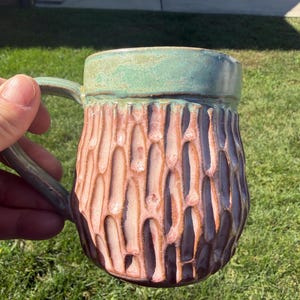 May include: A ceramic mug featuring a textured, carved exterior with a vertical pattern. The upper rim is glazed in a soft green, while the body transitions from a light pinkish-tan to a deep, dark purple at the base, creating a gradient effect.