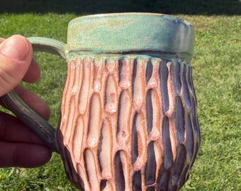 Pode incluir: Uma caneca de cerâmica com um exterior texturizado e esculpido com um padrão vertical. A borda superior é esmaltada num verde suave, enquanto o corpo transita de um rosa-bege claro para um roxo escuro profundo na base, criando um gradiente.
