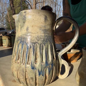 May include: A large, handmade ceramic pitcher with a curved handle. The pitcher has a beige and blue glaze with vertical, textured grooves around the base. The pitcher is sitting on a table outside.