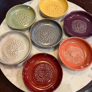 Pode incluir: Uma cole&ccedil;&atilde;o de sete pratos raladores circulares de cer&acirc;mica em v&aacute;rias cores, incluindo amarelo, verde, roxo, azul, branco, laranja e vermelho. Cada prato apresenta um padr&atilde;o central texturizado em relevo para ralar e pequenos orif&iacute;cios na borda.