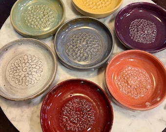 May include: A collection of seven circular ceramic grater plates in various colors including yellow, green, purple, blue, white, orange, and red. Each plate features a raised, textured center pattern designed for grating and small holes along the rim.