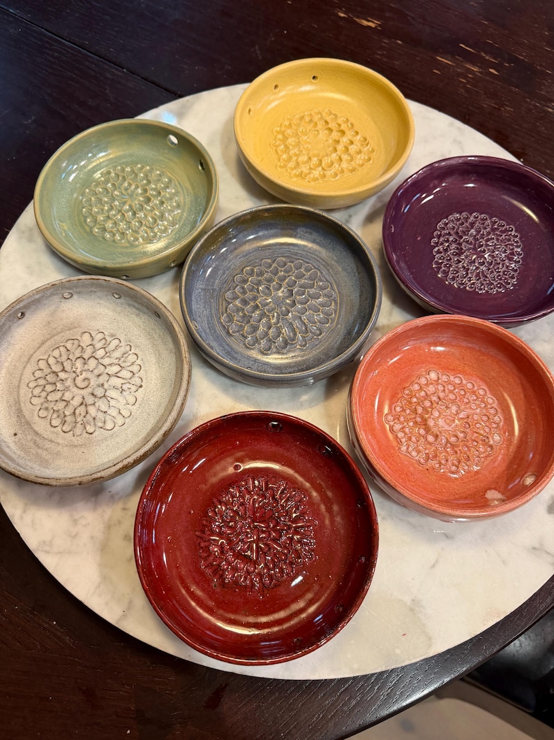 Pode incluir: Uma cole&ccedil;&atilde;o de sete pratos raladores circulares de cer&acirc;mica em v&aacute;rias cores, incluindo amarelo, verde, roxo, azul, branco, laranja e vermelho. Cada prato apresenta um padr&atilde;o central texturizado em relevo para ralar e pequenos orif&iacute;cios na borda.
