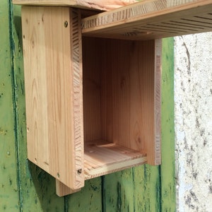 Puede incluir: Una casita de p&aacute;jaros de madera con un techo abatible y una peque&ntilde;a abertura para que los p&aacute;jaros puedan entrar. La casita de p&aacute;jaros est&aacute; hecha de madera natural y tiene un color marr&oacute;n claro.