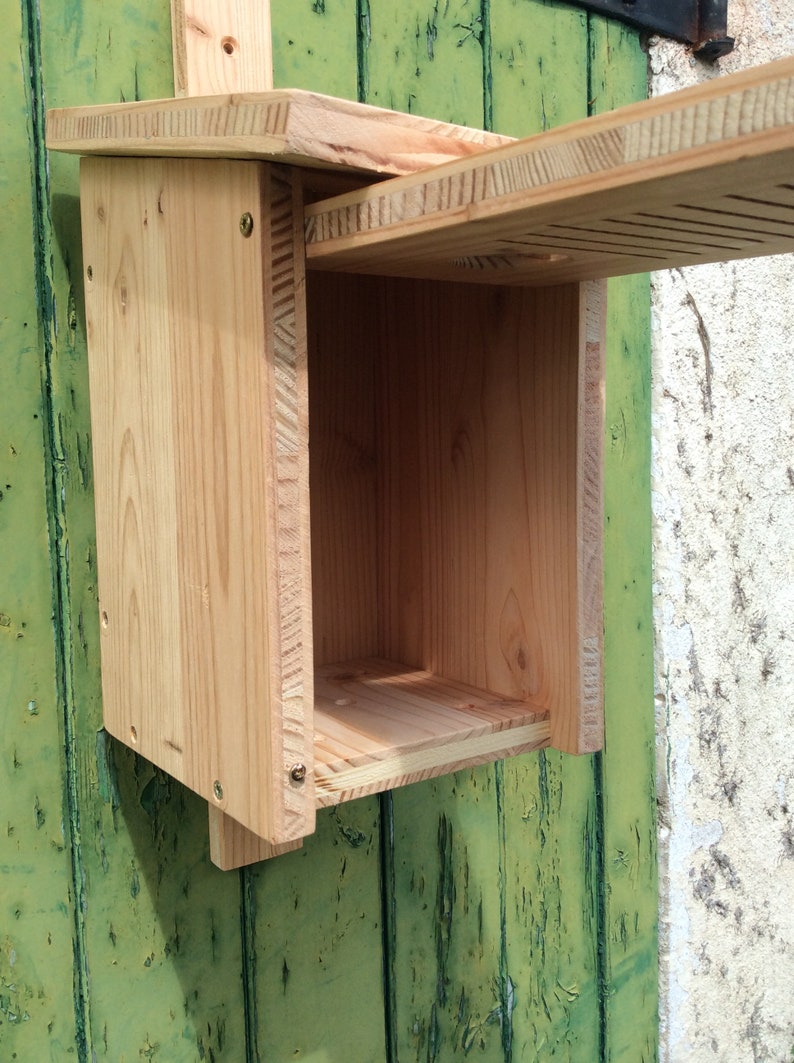 Puede incluir: Una casita de p&aacute;jaros de madera con un techo abatible y una peque&ntilde;a abertura para que los p&aacute;jaros puedan entrar. La casita de p&aacute;jaros est&aacute; hecha de madera natural y tiene un color marr&oacute;n claro.