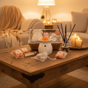 May include: A white ceramic oil burner with an orange flower, a woven basket containing various home fragrance items, a glass reed diffuser with black sticks, two small candles in glass jars, and packages of orange wax melts on a wooden table.