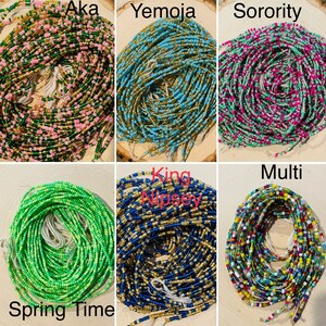 May include: An assortment of colourful beaded waist beads, including green, pink, blue, gold, and multi-coloured beads. The beads are arranged in a circular pattern and are labelled with names such as "Aka", "Yemoja", "Sorority", "King Nipsey", "Spring Time", and "Multi".