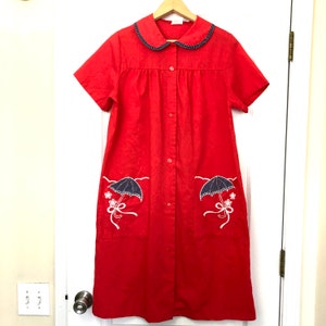 Könnte beinhalten: Ein roter, knopfverschluss-Hausmantel mit kurzen Ärmeln und einem weißen Kragen, der mit blauen Punkten besetzt ist. Der Hausmantel hat zwei Taschen mit gestickten weißen Regenschirmen und Blumen.