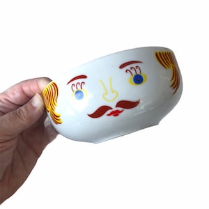 May include: White ceramic bowl with a whimsical face design. The bowl features a cartoon face with blue eyes, a red mustache, a yellow nose, and red and yellow hair. The bowl is held by a hand.