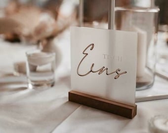 May include: White acrylic table number sign with the word "Tisch Eins" in a cursive font. The sign is on a wooden stand.
