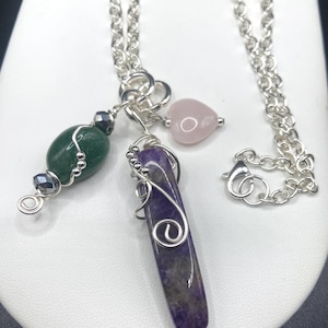 May include: A silver chain necklace with three gemstone pendants. The pendants include a green oval, a purple rectangular stone, and a pink heart-shaped stone. The stones are wire-wrapped and connected to the chain.