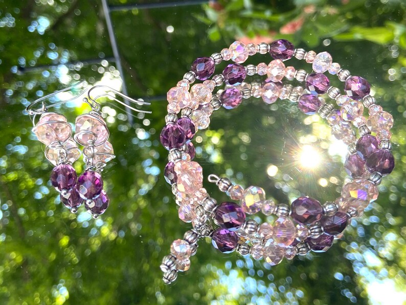 May include: A set of purple and pink crystal earrings and a matching bracelet. The earrings are dangle style and the bracelet is a wrap style.