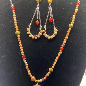May include: A necklace and earring set featuring a chain and wire design. The necklace has a pendant with a flower design and a mix of orange, yellow, and red beads. The earrings have a teardrop shape with matching beads.
