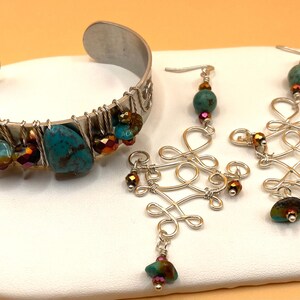 May include: A silver-toned metal cuff bracelet and matching earrings. The bracelet features turquoise stones and iridescent beads. The earrings have a wire design with turquoise and iridescent accents. The jewelry is displayed on a white surface.
