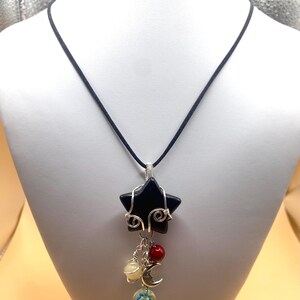 May include: A black star-shaped pendant necklace with silver wire detailing. The pendant hangs from a black cord and features dangling charms, including a red bead, a yellow bead, a crescent moon, and a glass bead with a floral design.