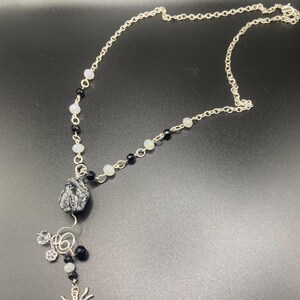 May include: Silver-toned necklace featuring a spider charm, black and white beads, and a speckled stone pendant. The necklace has a delicate chain and a unique, handcrafted design, perfect for adding a touch of gothic flair to any outfit.