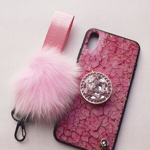 May include: A pink phone case with a cracked pink and gold design. The case has a clear rhinestone ring holder and a pink fluffy keychain with a silver clip.